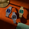 Three Casio retro wristwatches with metal link bands are arranged on a textured orange surface. One watch has a blue dial and silver case, one has a black dial and silver case, and one has a green dial with a gold case.
